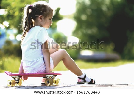 Pretty cute little blond girl in white shorts and T-shirt sitting on pink skateboard and smiling in camera on a light blurred summer background. Children activities and active lifestyle concept.