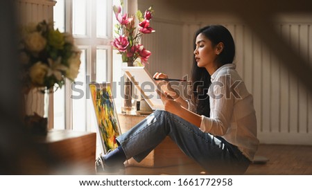 Photo of young artist girl holding a paint brush and drawing an oil colors on canvas while sitting at the modern arts studio. Concept of creative woman as artist.