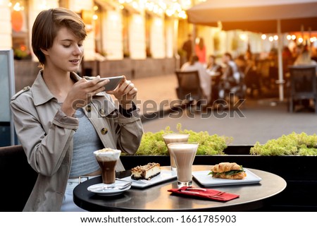 Pretty young girl teen blogger cafe table modern smart phone take post photo food coffee dessert instagram feed shoot vlog for restaurant review camera romantic cafeteria terrace background copyspace.