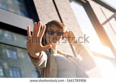 Teen hipster short hair gen z girl wear stylish glasses stand on urban sunny city street showing stop hand gesture. Female igen teenage fashion model woman look at camera outdoor, portrait.
