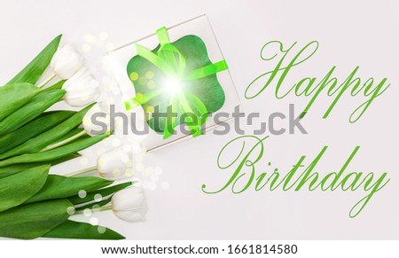 Happy Birthday text message sign on light background. White tulip flowers and gift box with green ribbon bow flat lay. Flower Bouquet congratulations greeting card. Copy space website banner top view.