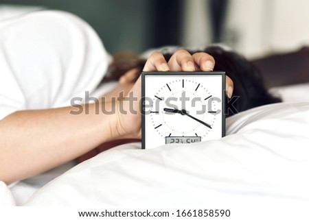 Sleepy girl's hand showing alarm clock and waking up late in the morning, Woman sleep in and laying down in bed hiding under the blanket, Sleep disorder and not enough rest problem.