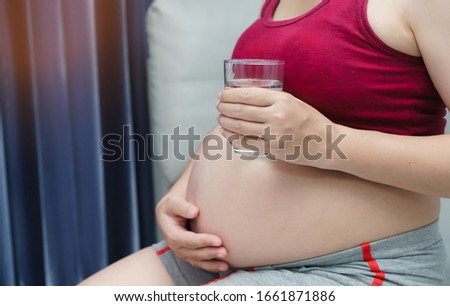 Close-up pregnant woman holding  glass of water sitting on sofa in the room. Nutrition and diet during pregnancy.