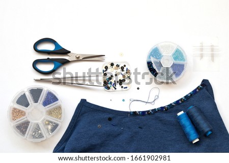 Needlework and beadwork. Decoration of a blue top with bugle embroidery, bead and sequins. Accessories for embroidery: sets of beads in boxes, tweezers, needles and scissors on a white background Royalty-Free Stock Photo #1661902981