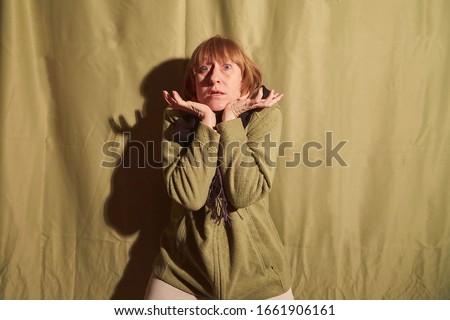 A middle-aged woman poses showing clothes near a crumpled green curtain. An inept model in non-professional shooting. Photography for sales cardigan on the Internet or online store