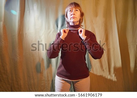 A middle-aged woman poses showing clothes near a crumpled green curtain. An inept model in non-professional shooting. Photography for turtleneck sales on the Internet or online store