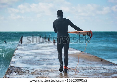 Surfer is walking on pier and getting ready to jump into the waves and enjoying surfing during windy and cloudy day.