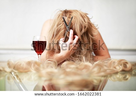 Blonde adult caucasian woman cutting her hair with scissors and drink glass of red wine