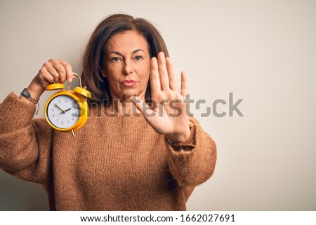 Middle age brunette woman holding clasic alarm clock over isolated background doing stop sing with palm of the hand. Warning expression with negative and serious gesture on the face.