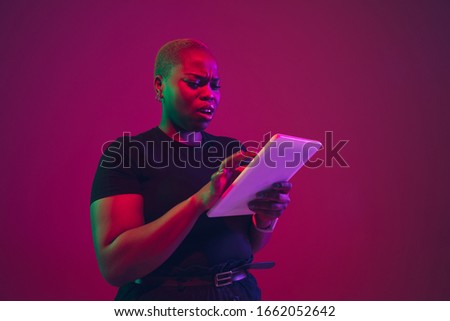 Using tablet. African-american young woman's portrait on purple background. Beautiful model in black shirt. Concept of emotions, facial expression, sales, ad, inclusion, diversity. Copyspace.