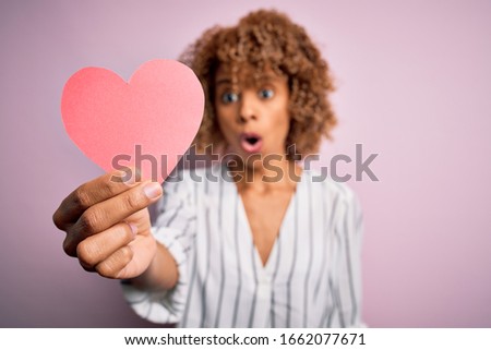 Young african american romantic woman with curly hair holding paper heart shape scared in shock with a surprise face, afraid and excited with fear expression