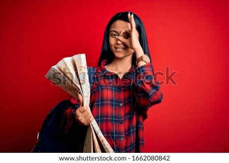 Young beautiful chinese tourist woman holding city map over isolated red background with happy face smiling doing ok sign with hand on eye looking through fingers
