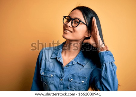 Young beautiful chinese woman wearing casual denim shirt over isolated yellow background smiling with hand over ear listening an hearing to rumor or gossip. Deafness concept.