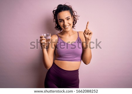Young beautiful sportswoman with curly hair doing sport drinking glass of water surprised with an idea or question pointing finger with happy face, number one