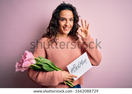 Beautiful curly hair woman holding best mom message and tulips celebrating mothers day doing ok sign with fingers, excellent symbol