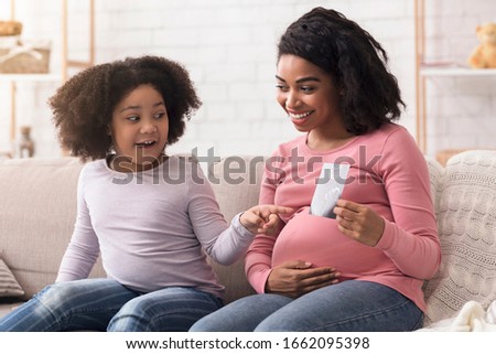 Pregnant african american mom showing ultrasound scan photo to her excited little daughter, cute girl pointing at picture with amazement