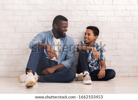 Men Family. Portrait Of Happy African American Father And Son Sitting Together On Floor Against White Brick Wall And Lookig At Ech Other, Copy Space Royalty-Free Stock Photo #1662103453