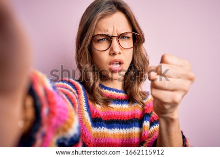 Young blonde girl wearing glasses taking a selfie picture of herself over pink isolated background annoyed and frustrated shouting with anger, crazy and yelling with raised hand, anger concept