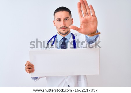 Young doctor man holding blank advertising banner over isolated background with open hand doing stop sign with serious and confident expression, defense gesture