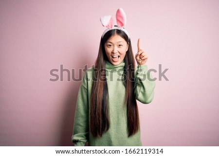 Young asian woman wearing cute easter bunny ears over pink background pointing finger up with successful idea. Exited and happy. Number one.