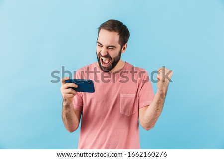 Image of young emotional excited screaming man isolated over blue wall background play games by mobile phone make winner gesture.