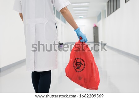 Scientist wearing blue gloves and red bag with public bioharzard sign.A woman worker hand holding red garbage bag.Maid and infection waste bin at the indoor public building.Infectious control. Royalty-Free Stock Photo #1662209569