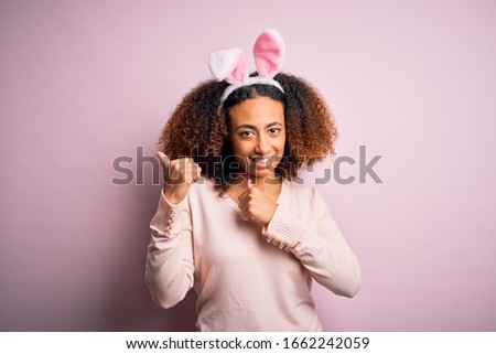 Young african american woman with afro hair wearing bunny ears over pink background Pointing to the back behind with hand and thumbs up, smiling confident