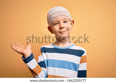 Young little caucasian kid injured wearing medical bandage on head over yellow background smiling cheerful presenting and pointing with palm of hand looking at the camera.