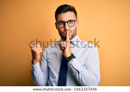 Young handsome businessman wearing tie and glasses standing over yellow background asking to be quiet with finger on lips pointing with hand to the side. Silence and secret concept.