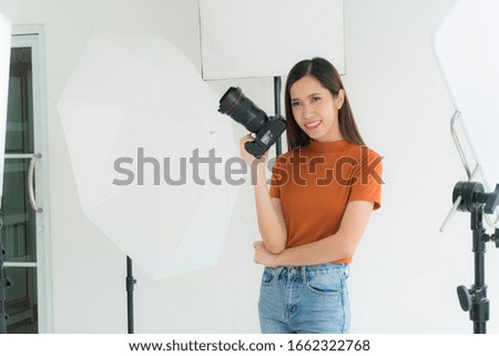 Asian photographer is picking up a digital camera to take pictures in her studio with lighting equipment on background. Photography studio concept
