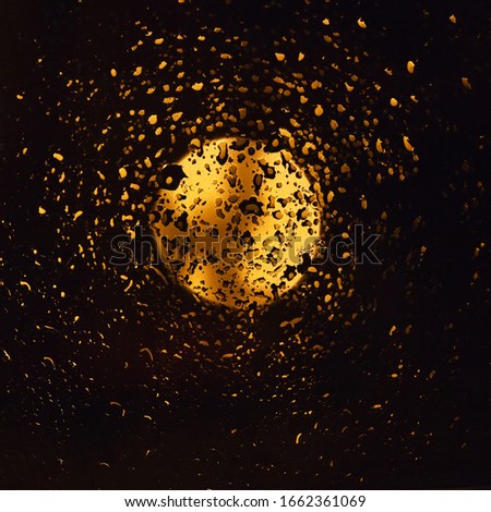 Drops of rain and light of the full moon on dark glass at night. Wet window glass with lots of rain drops and a view of the night blac sky with the yellow gold glowing moon. Abstract background