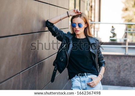 Blonde girl wearing black t-shirt, glasses and leather jacket posing against street , urban clothing style. Street photography