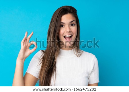 Teenager girl over isolated blue background surprised and showing ok sign