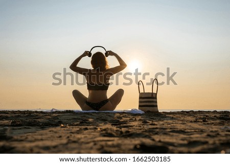 Young yoga woman in the bikini (swimsuit) and headphones meditation on the ocean beach at sunrise. 
