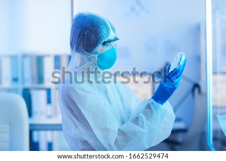 Horizontal side view shot of unrecognizable medical laboratory scientist holding petri dish with virus specimen looking at it