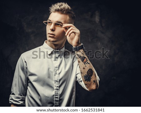 Interested and inked hipster male model posing in a studio, holding his glasses, wearing a white shirt, standing in front of the grey background, looking cool and interested