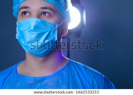close-up portrait of a caucasian doctor surgeon, in a sterile suit, mask, holds a lamp, pointing it to the camera. On a blue background. Operating room. Surgery.