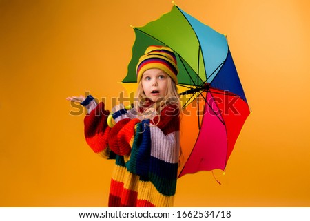 child girl smiling in a bright multicolored sweater and hat holding an umbrella stands on a yellow background. little girl in bright clothes is holding a red-colored umbrella. happy child, space for