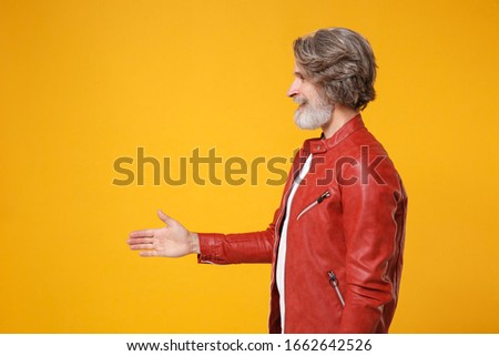 Side view of elderly gray-haired mustache bearded man in red leather jacket isolated on yellow background. People lifestyle concept. Mock up copy space. Standing with outstretched hand for greeting