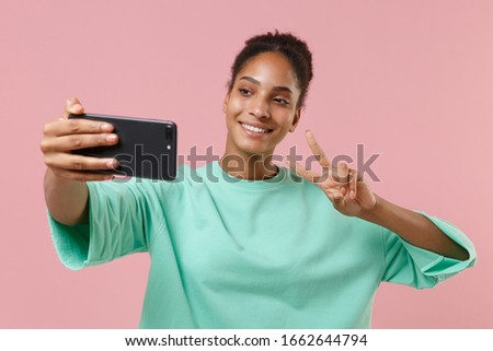 Smiling young african american woman girl in green sweatshirt isolated on pastel pink background. People lifestyle concept. Mock up copy space. Doing selfie shot on mobile phone, showing victory sign