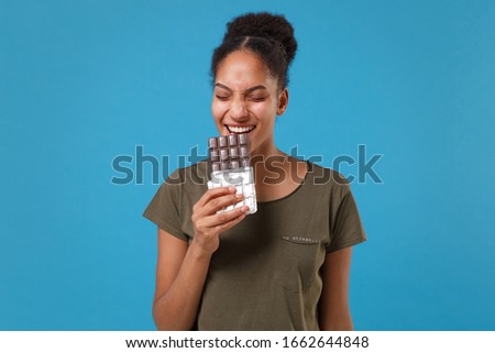 Funny young african american woman girl in casual t-shirt posing isolated on bright blue background studio portrait. People emotions lifestyle concept. Mock up copy space. Biting eating chocolate bar