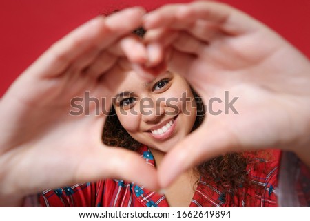 Smiling african american girl in pajamas homewear resting at home isolated on red background. Relax good mood lifestyle concept. Mock up copy space. Showing shape heart with hands, heart-shape sign
