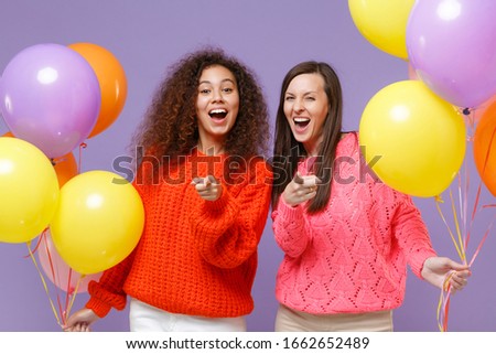 Laughing two european african american women friends in knitted sweaters isolated on violet purple background. Birthday holiday party concept. Celebrating hold colorful air balloons point on camera