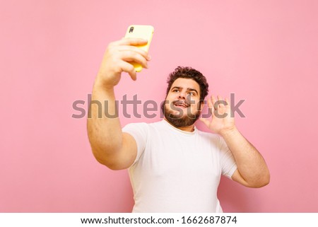 Funny fat man with a beard holds a smartphone in his hand and phone over a video call, isolated on a pink background.Overweight man communicates on video with a smile on his face. Copy space