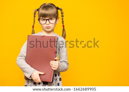 Little cheerful girl in glasses with serious face and funny pigtails holding school books against yellow background