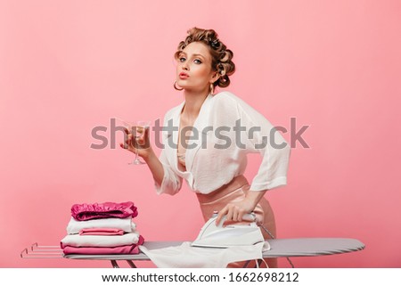 Woman in light silk outfit posing on pink background with martini glass and ironing clothes