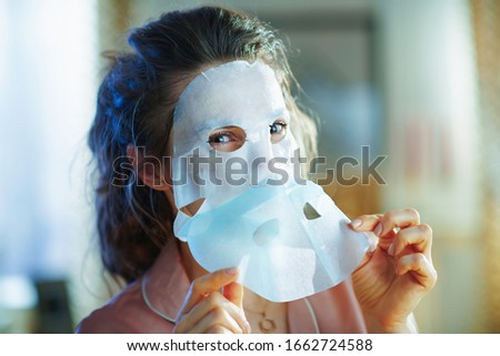 stylish 40 years old woman in pajamas at modern home in sunny winter day applying sheet facial mask and removing protecting layer.