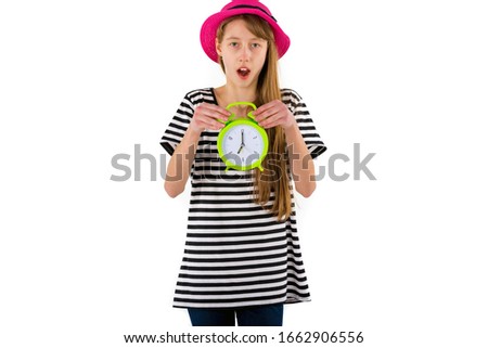 Surprised young caucasian girl wearing red hat dressed in white striped black  t-shirt holds alarm clock and looking at camera isolated over white background.Copy space banner advertisement.
