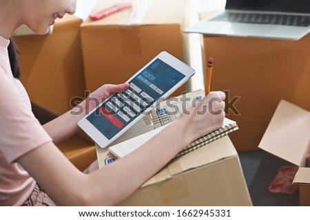 Photo of young beautiful woman packing her goods for delivery to customer by using computer tablet and laptop at the comfortable living room. Start up/Entrepreneur concept.