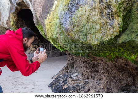 White man with a red jacket taking a very close photo with his smartphone of a rock in cathedral cove beach, Te Whanganui-A-Hei Marine Reserve, Coromandel Peninsula, north island, New Zealand 